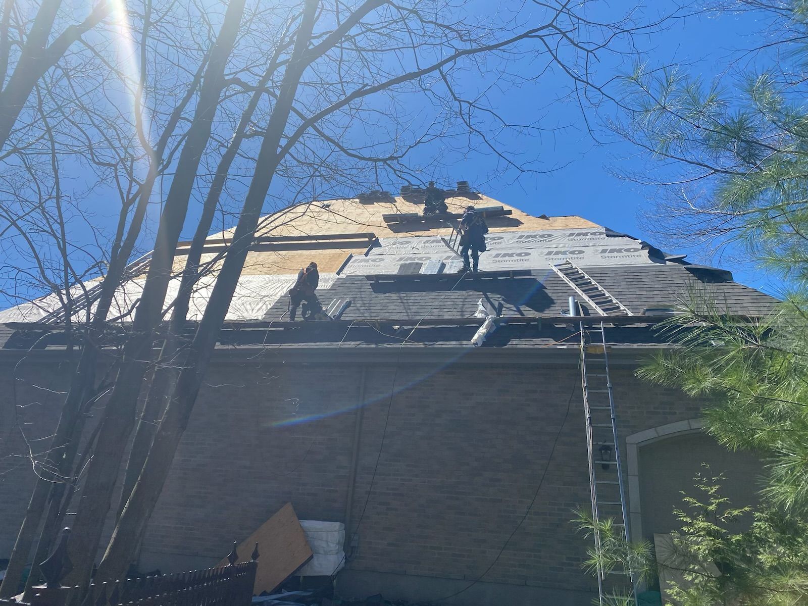 Des couvreurs travaillent sur un toit pentu, une échelle appuyée contre un bâtiment en briques sous un ciel bleu.