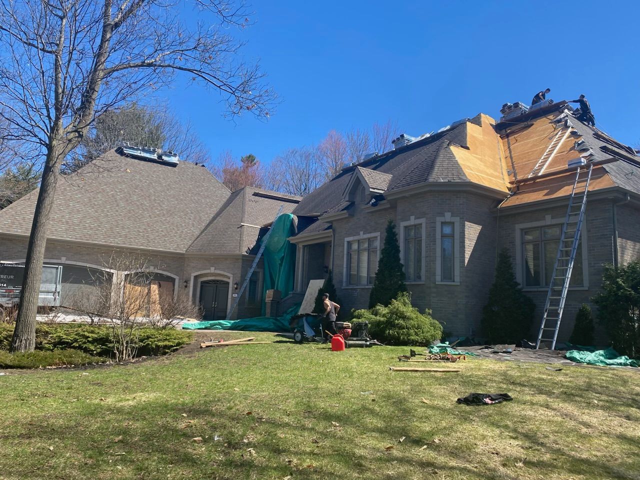 Grande maison aux toits gris et beiges, pelouse verte et une personne travaillant dans le jardin sous un ciel bleu azur.