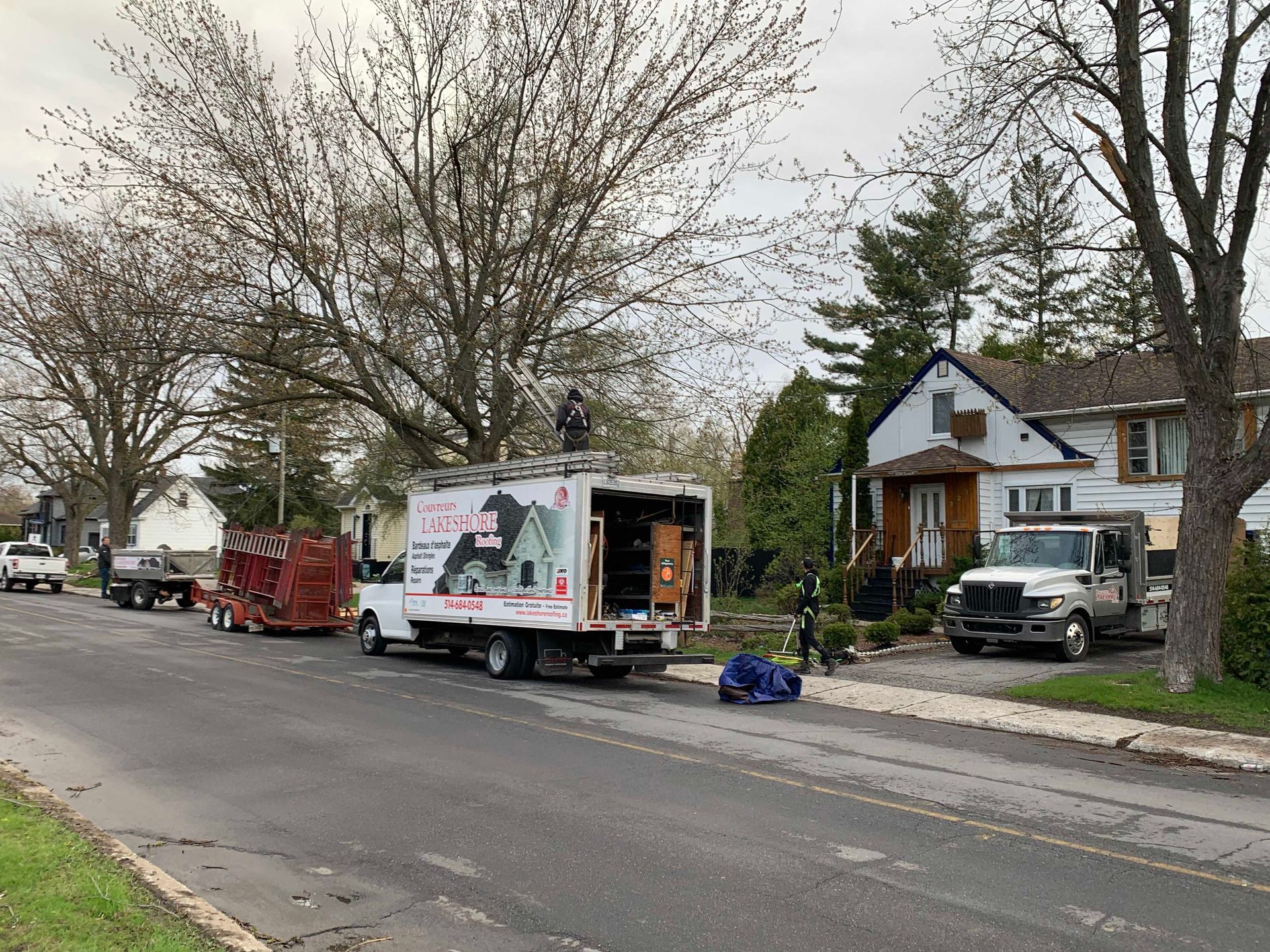 Street scene with two moving trucks, parked vehicles, and workers on a residential road with bare trees.