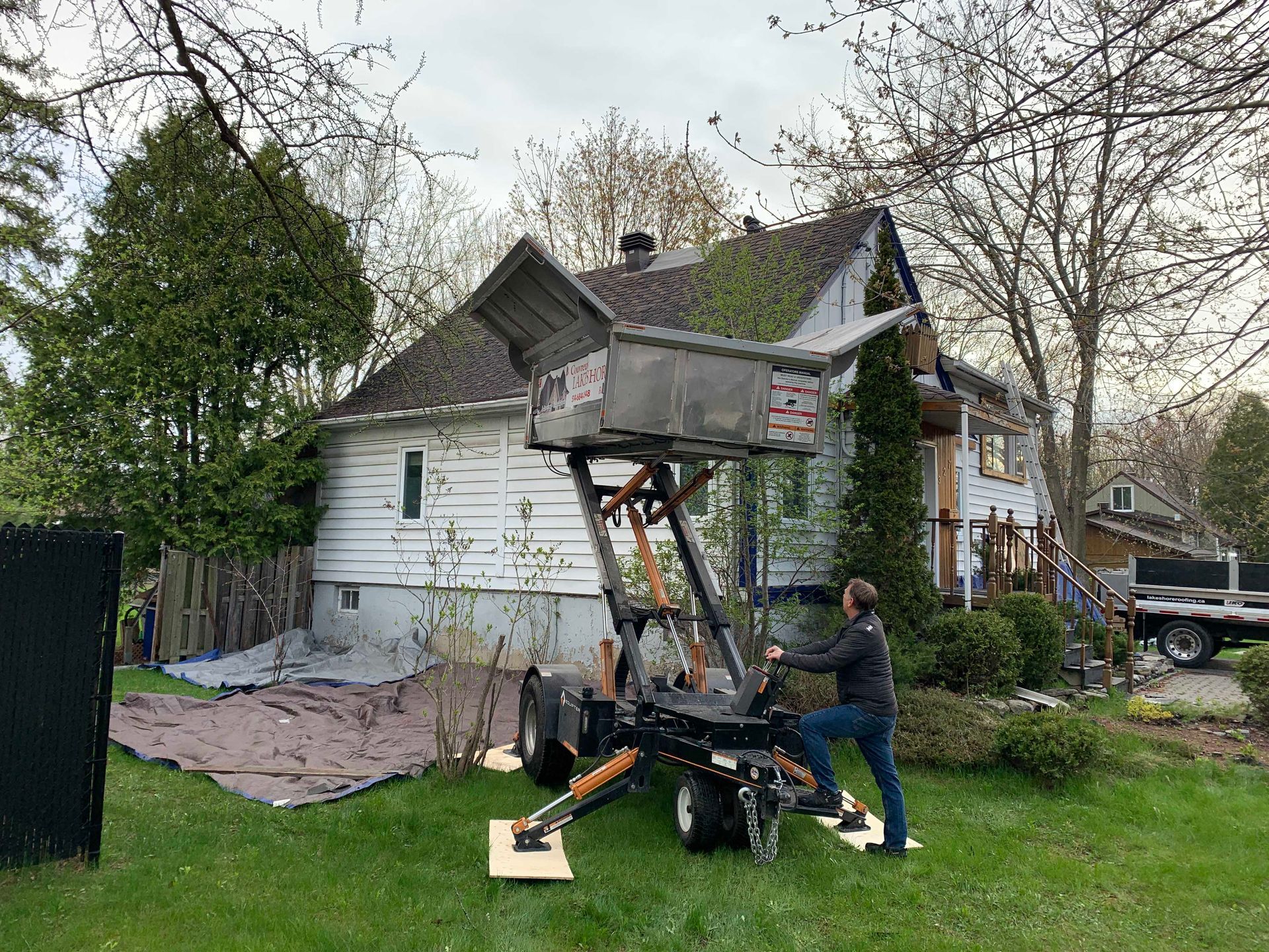 Une personne manœuvre une petite excavatrice à côté d'une maison blanche dont le revêtement et le toit sont endommagés.
