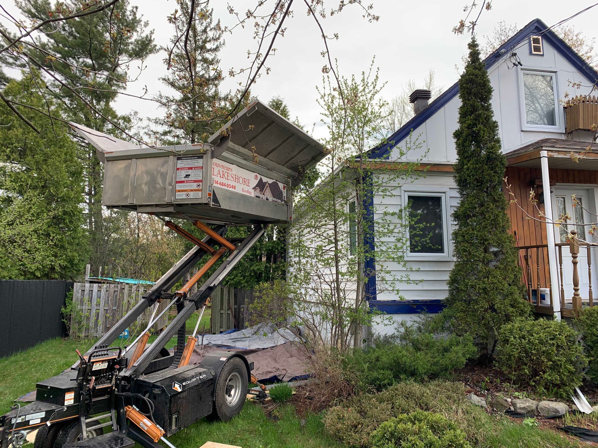 Un camion-benne à chargement frontal soulève une benne surélevée à côté d'une maison bleue et d'arbres