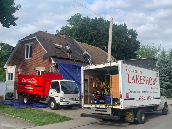 Two trucks parked at a house while workers repair the roof under a blue tarp.