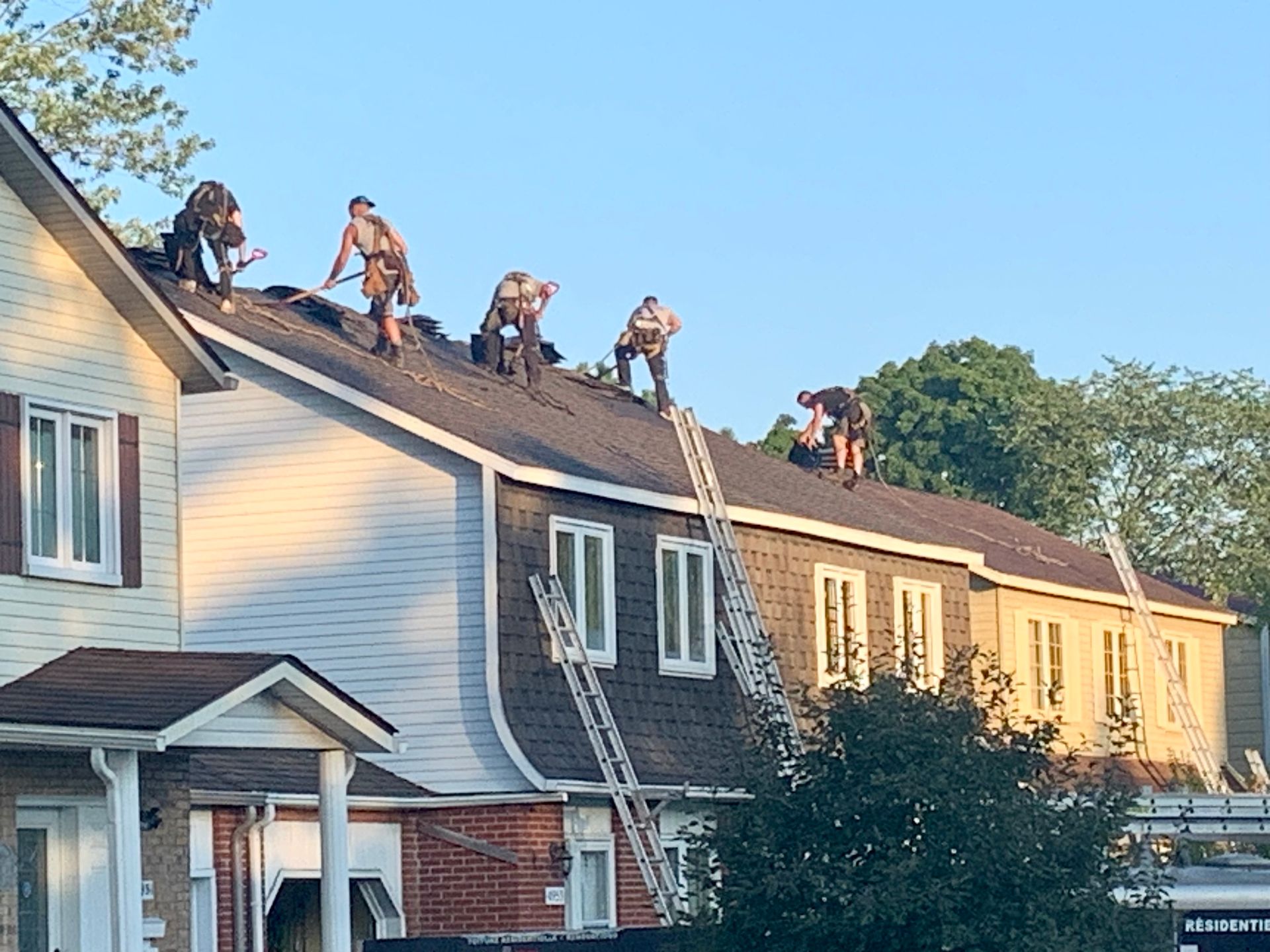 Des ouvriers réparent les bardeaux d'une rangée de maisons sur un toit, leurs échelles appuyées contre le bâtiment.
