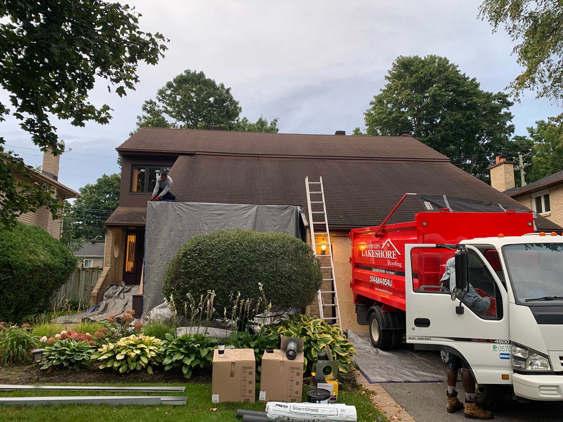 Des ouvriers refont le toit d'une maison ; le toit est recouvert d'une bâche et un camion utilitaire rouge est garé devant.