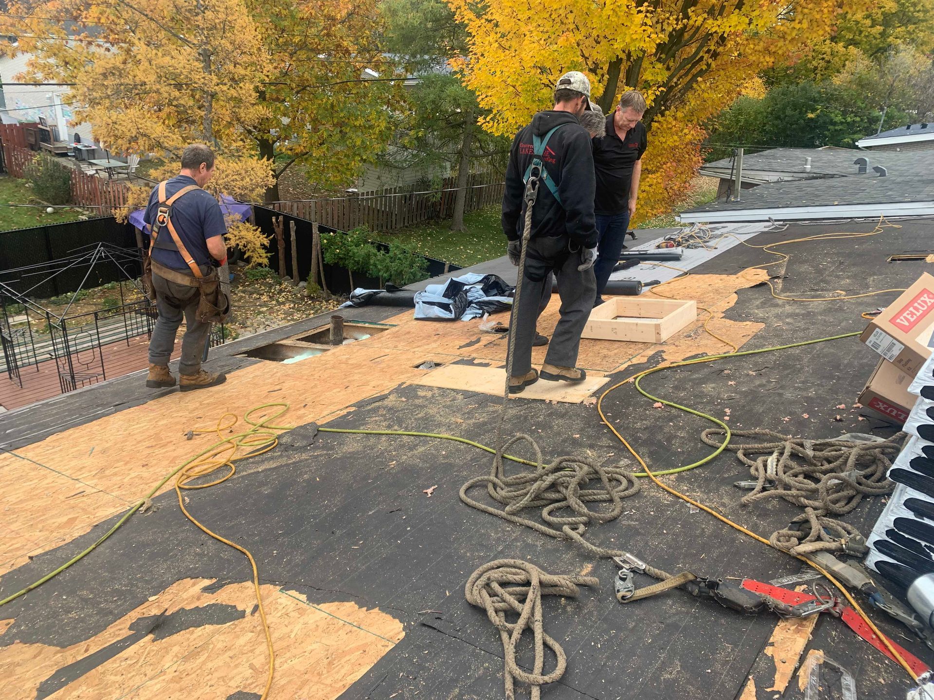Des couvreurs posent une sous-couche sur un toit en pente à l'aide de cordes et d'outils, avec des arbres d'automne en arrière-plan.
