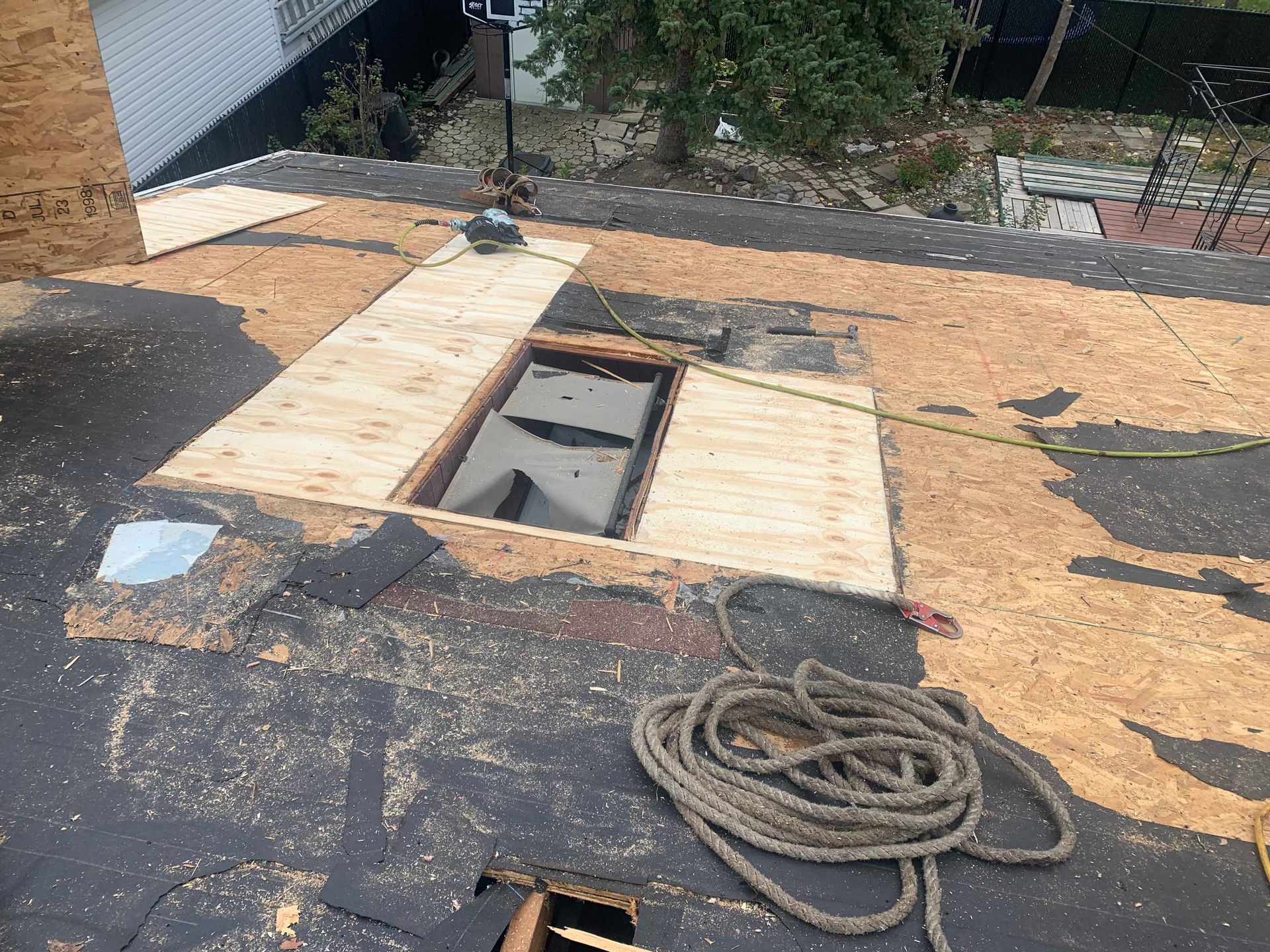 Toit avec une trappe ouverte recouverte de contreplaqué, toiture noire et une corde enroulée au premier plan.