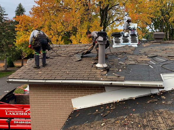 Roofers repairing storm-damaged shingle roof with exposed plywood and debris on a house