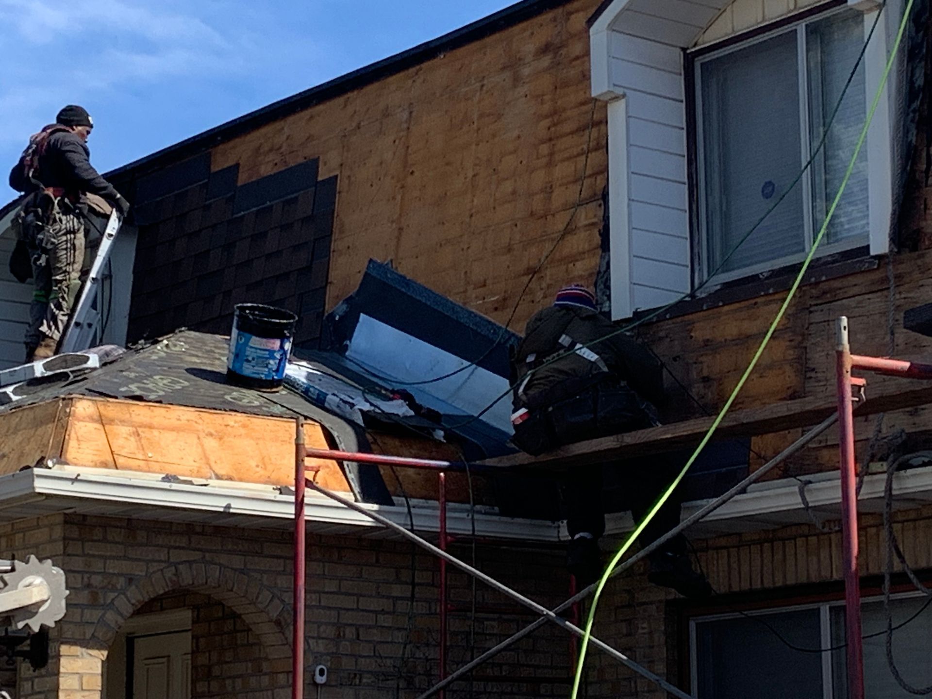 Ouvrier sur un échafaudage réparant un toit endommagé sur une maison en briques, avec des bardeaux et des débris apparents.