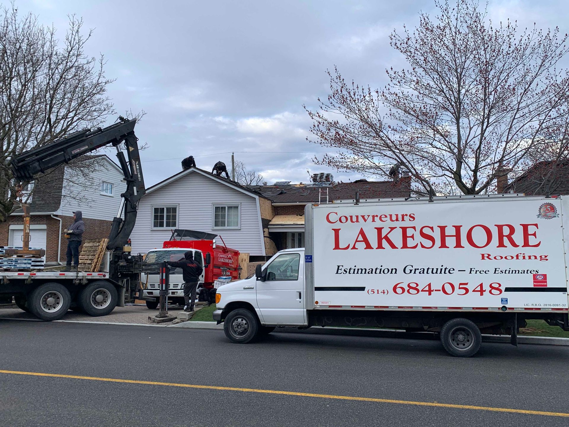 Un camion de déménagement Lakshore est garé à côté d'une grue devant une maison avec une voiture rouge et des arbres dénudés.