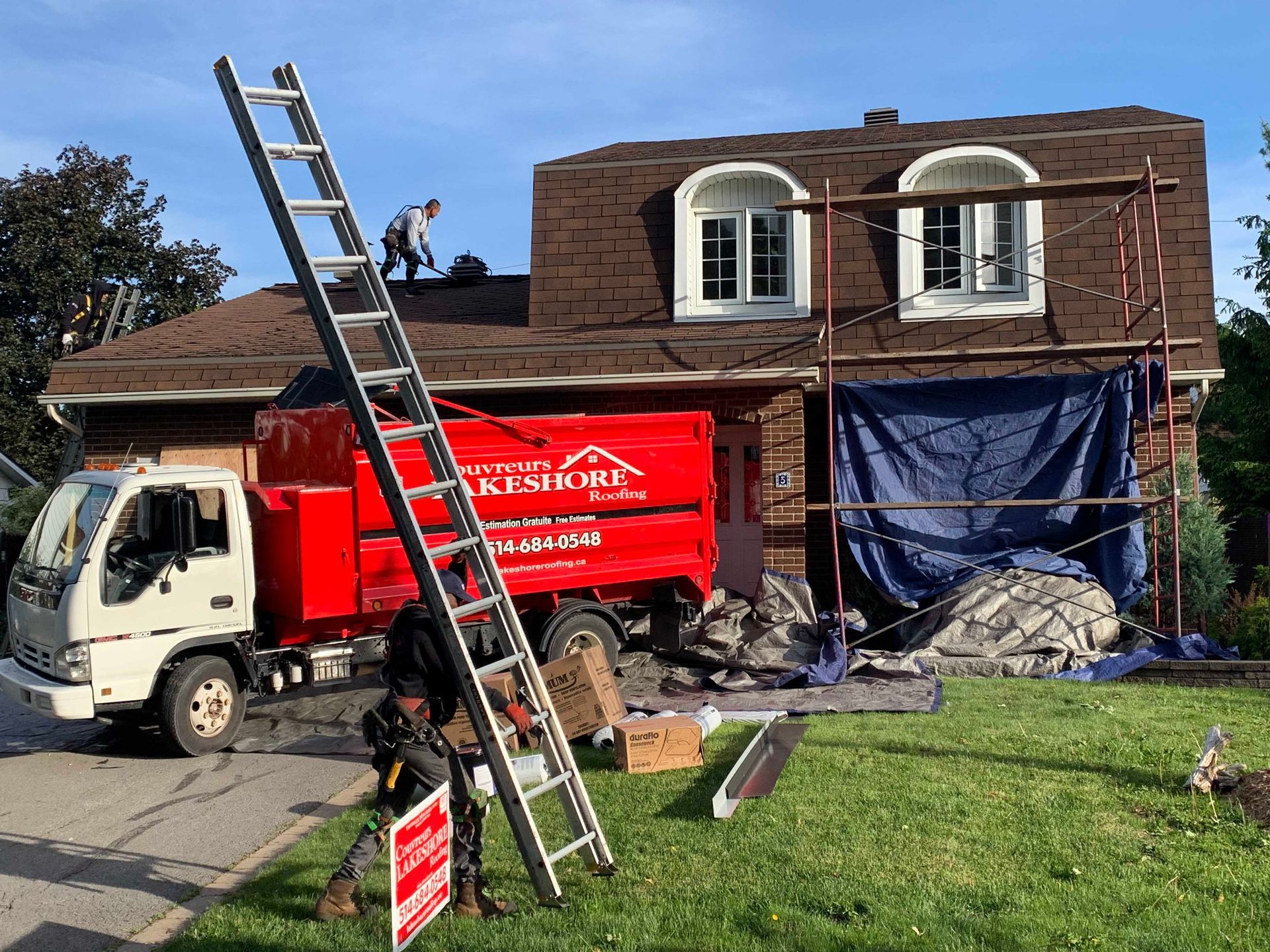 Camion-échelle et ouvriers devant une maison en briques avec échafaudages et bâche bleue sous un ciel nuageux