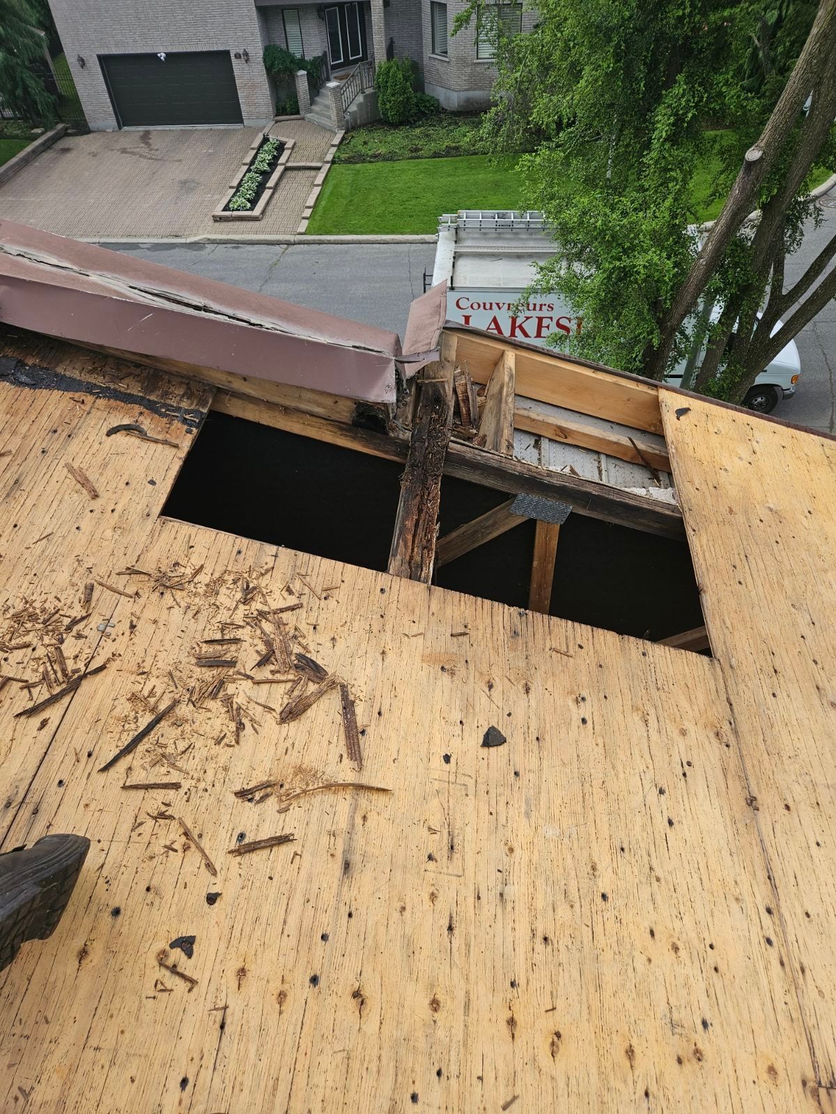 Toiture en contreplaqué endommagée, présentant de grands trous et des débris, donnant sur une rue résidentielle et une cour.