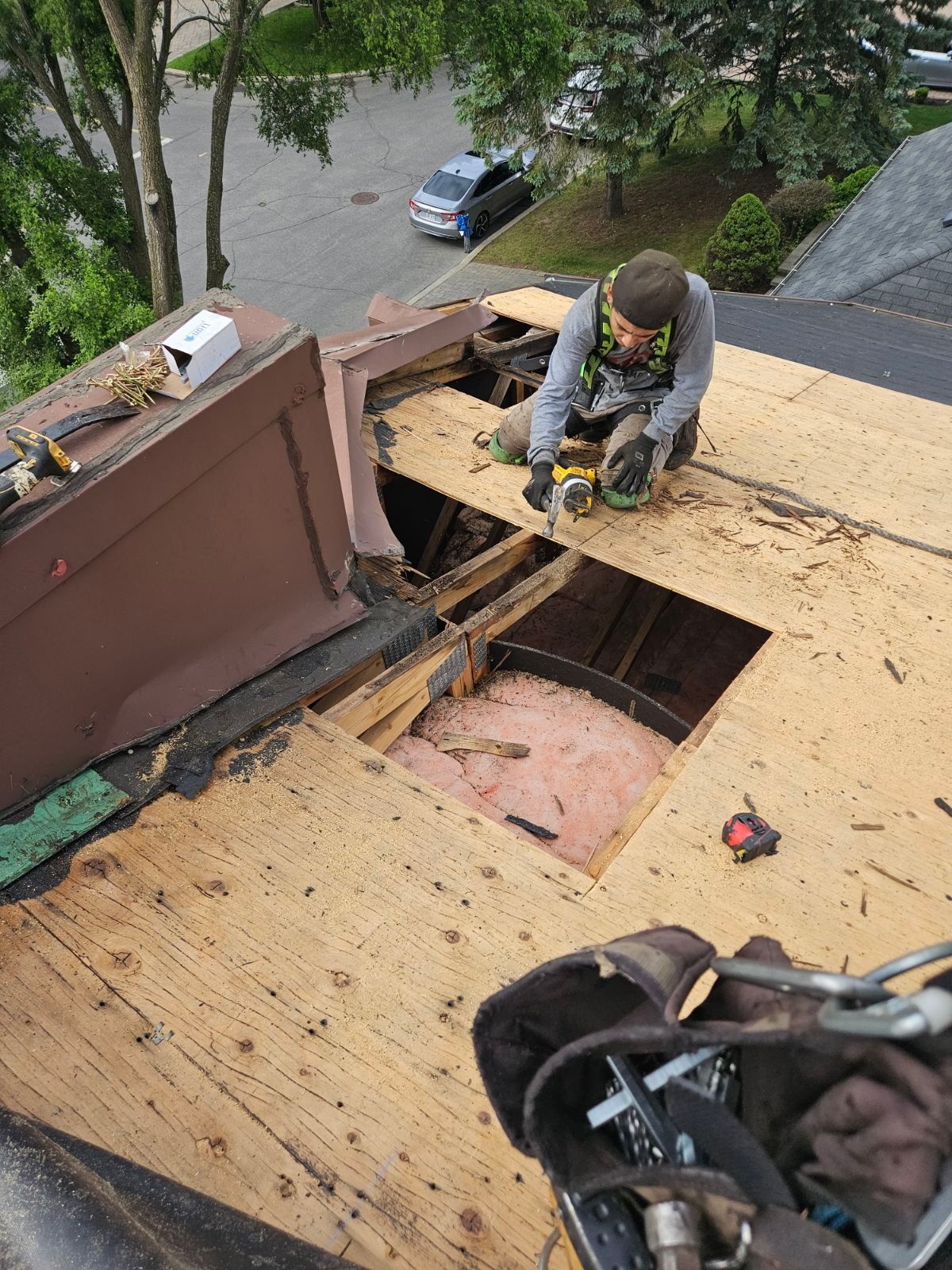Worker using a power tool on a roof with a large opening, near trees and a parked car below