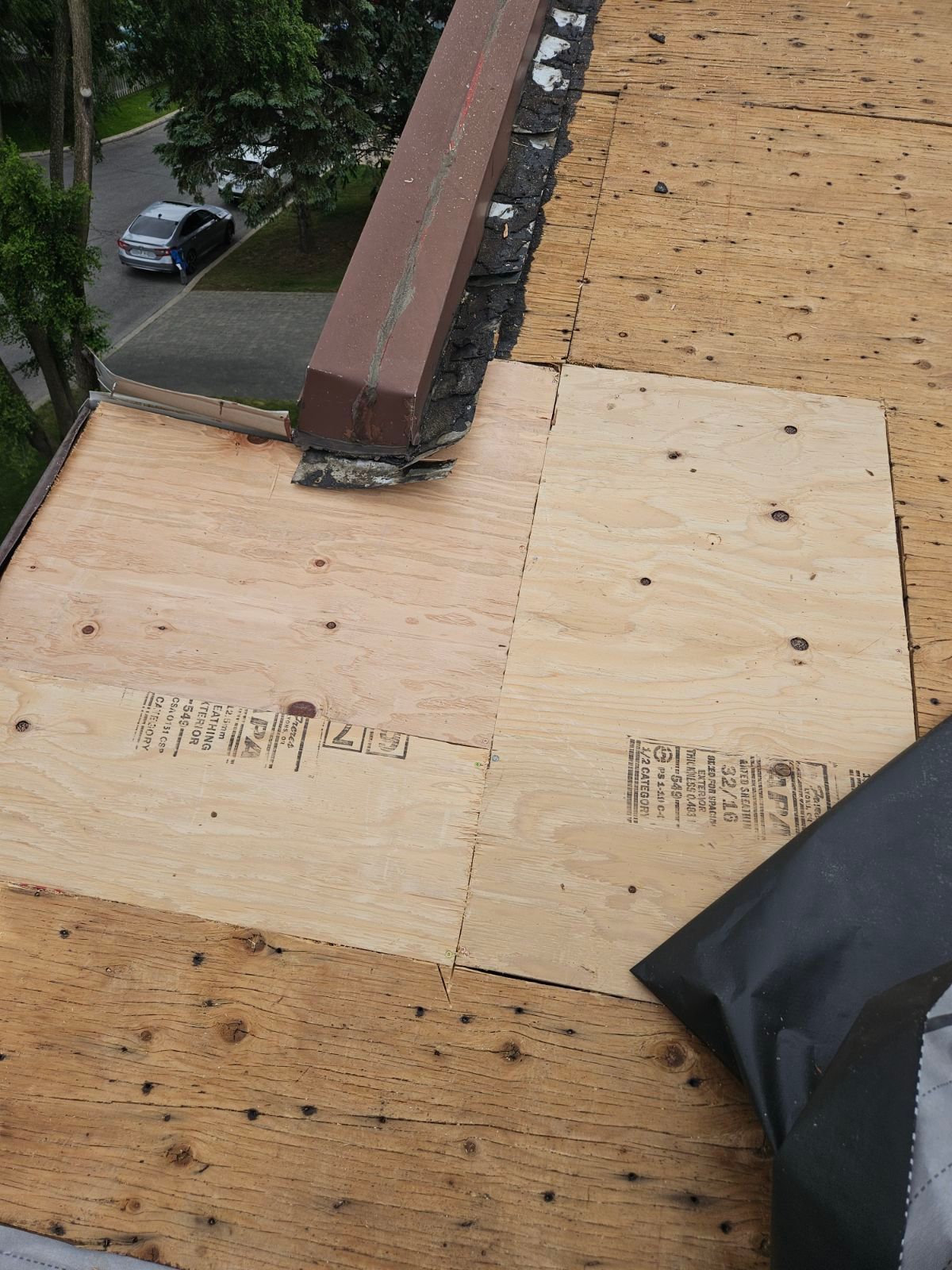 Réparation d'un toit en contreplaqué autour d'une cheminée en briques, avec un matériau de couverture noir et des arbres à proximité en contrebas.