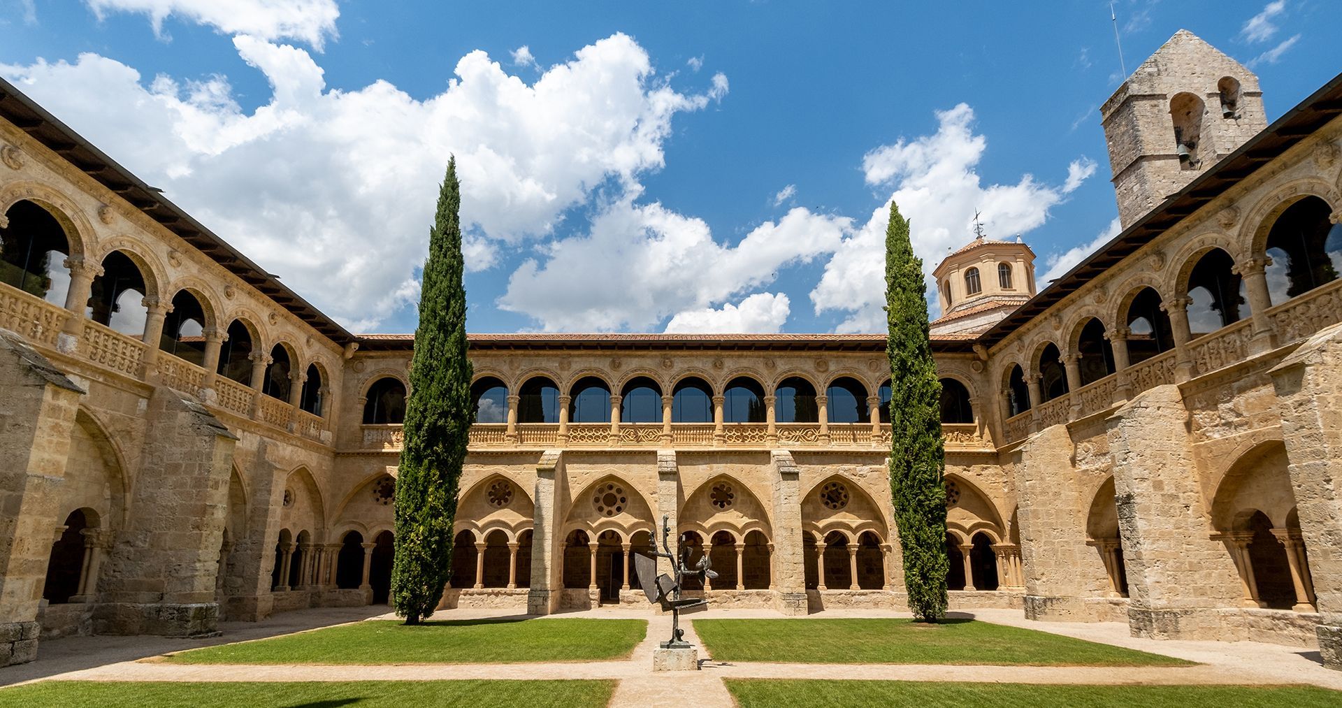 Courtyard of an old stone building with arched walkways, green lawn, and blue sky.
