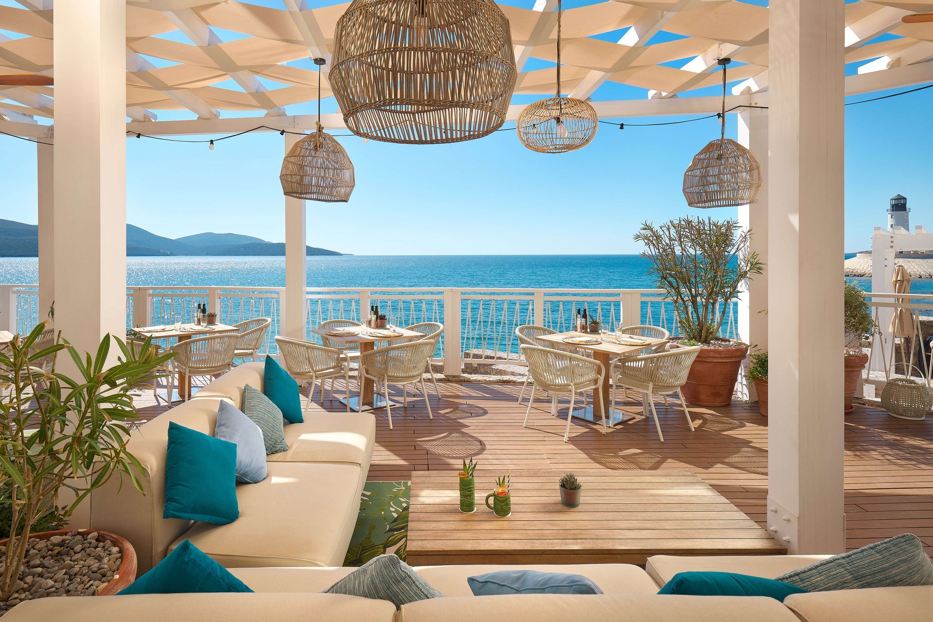 Outdoor restaurant overlooking the ocean with beige couches, tables, wicker lamps, and blue accents.