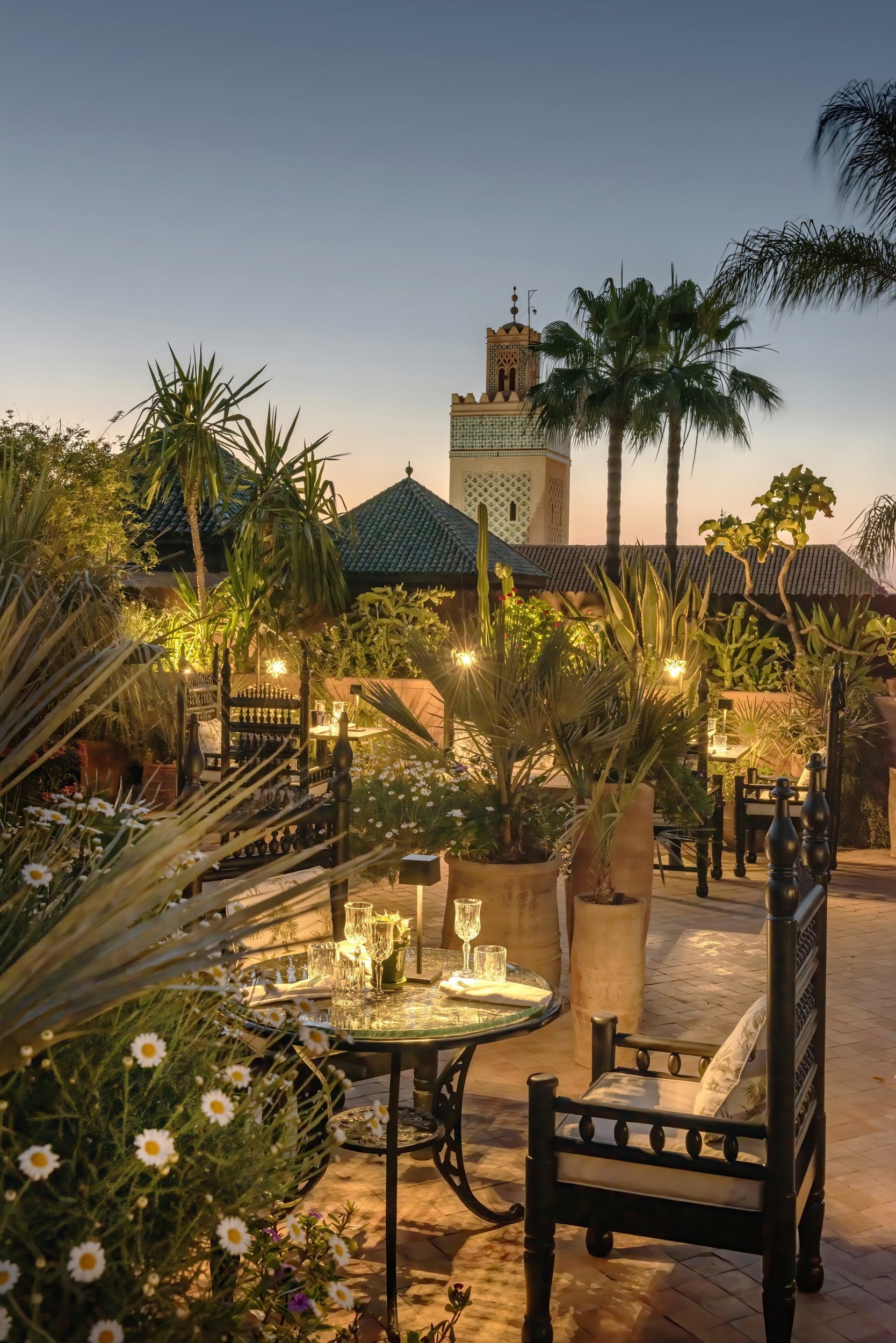Lantern-lit rooftop garden at dusk with palms, tables, and a glowing skyline view of Marrakech