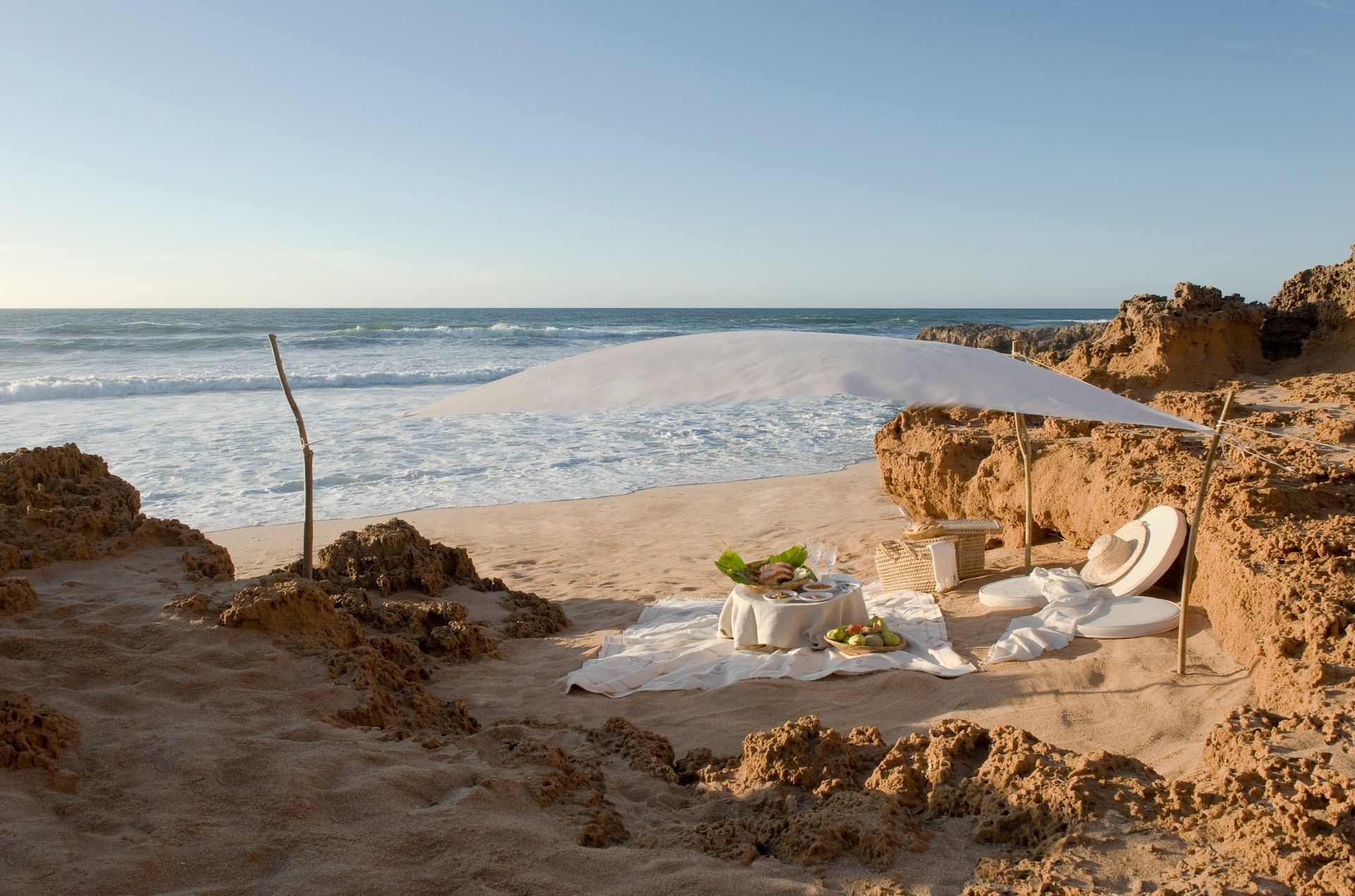 Beachside lounge chairs and table under a canopy beside rocky shore and calm ocean
