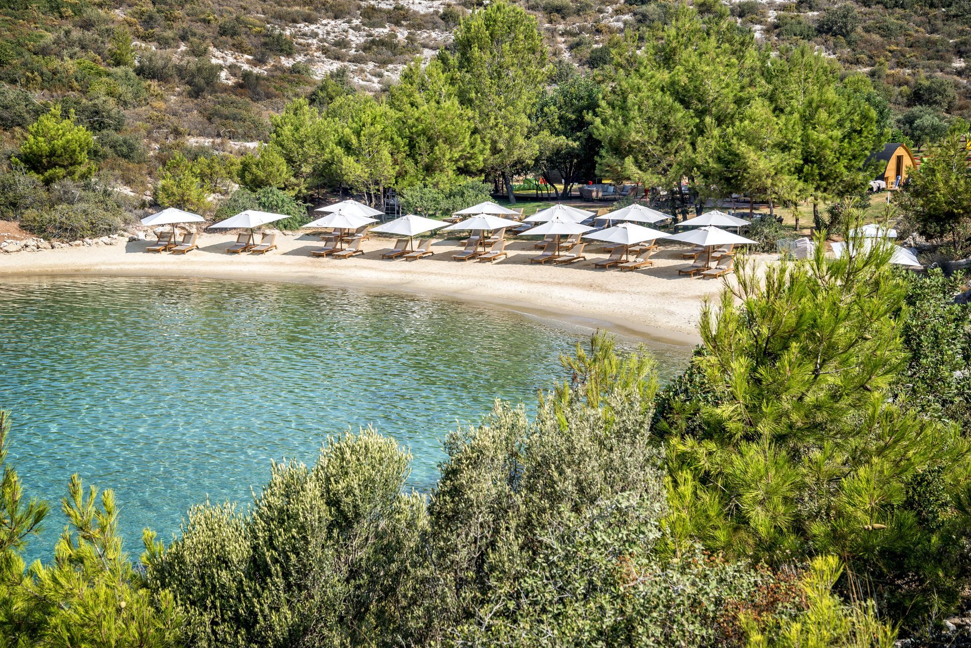 Beach with clear turquoise water, sandy shore, and white umbrellas. Lush green trees surround the cove.