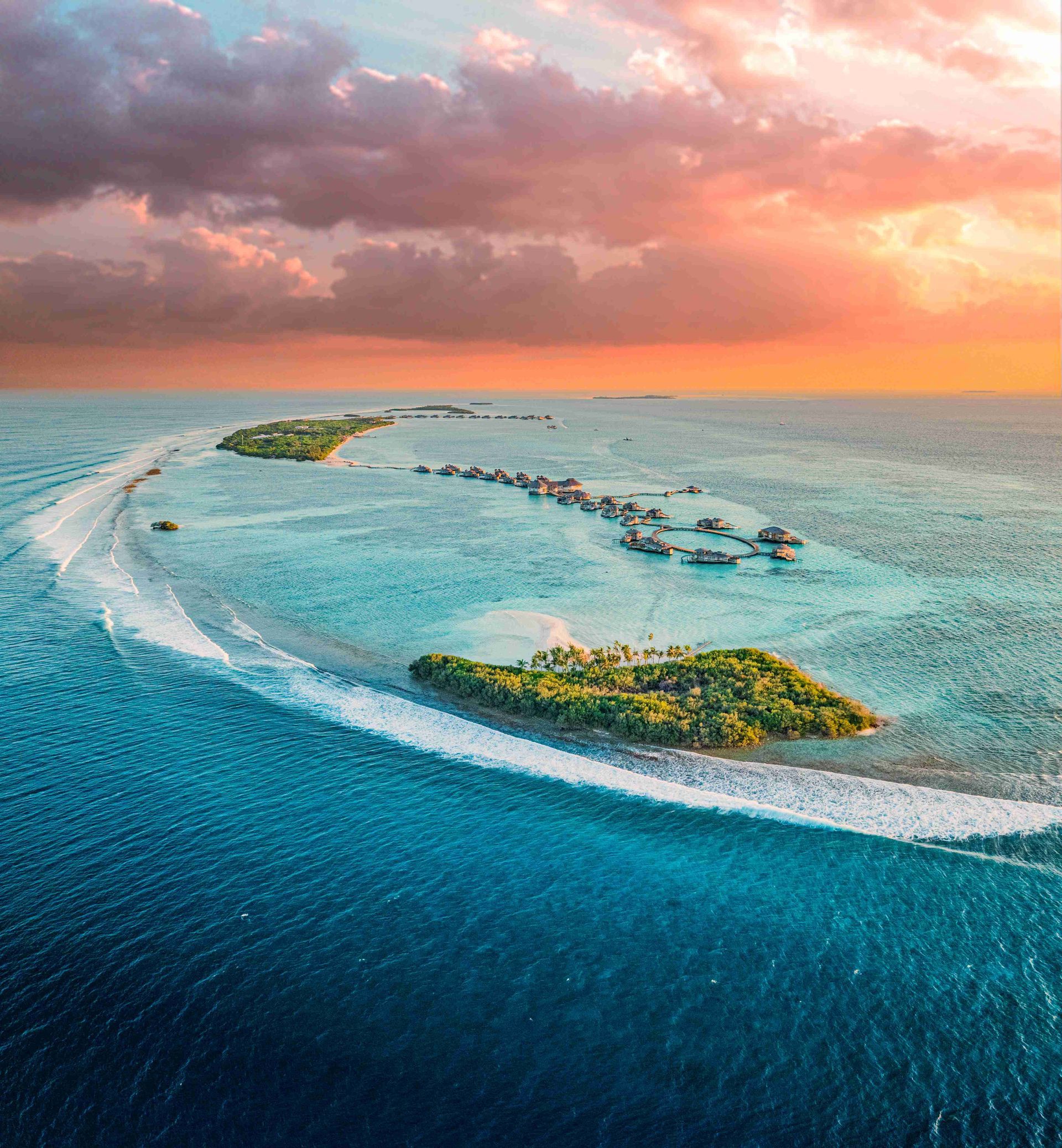 Aerial view of Soneva Jani's turquoise ocean, curved sandbar, and small eco islands under a pink-orange sunset sky
