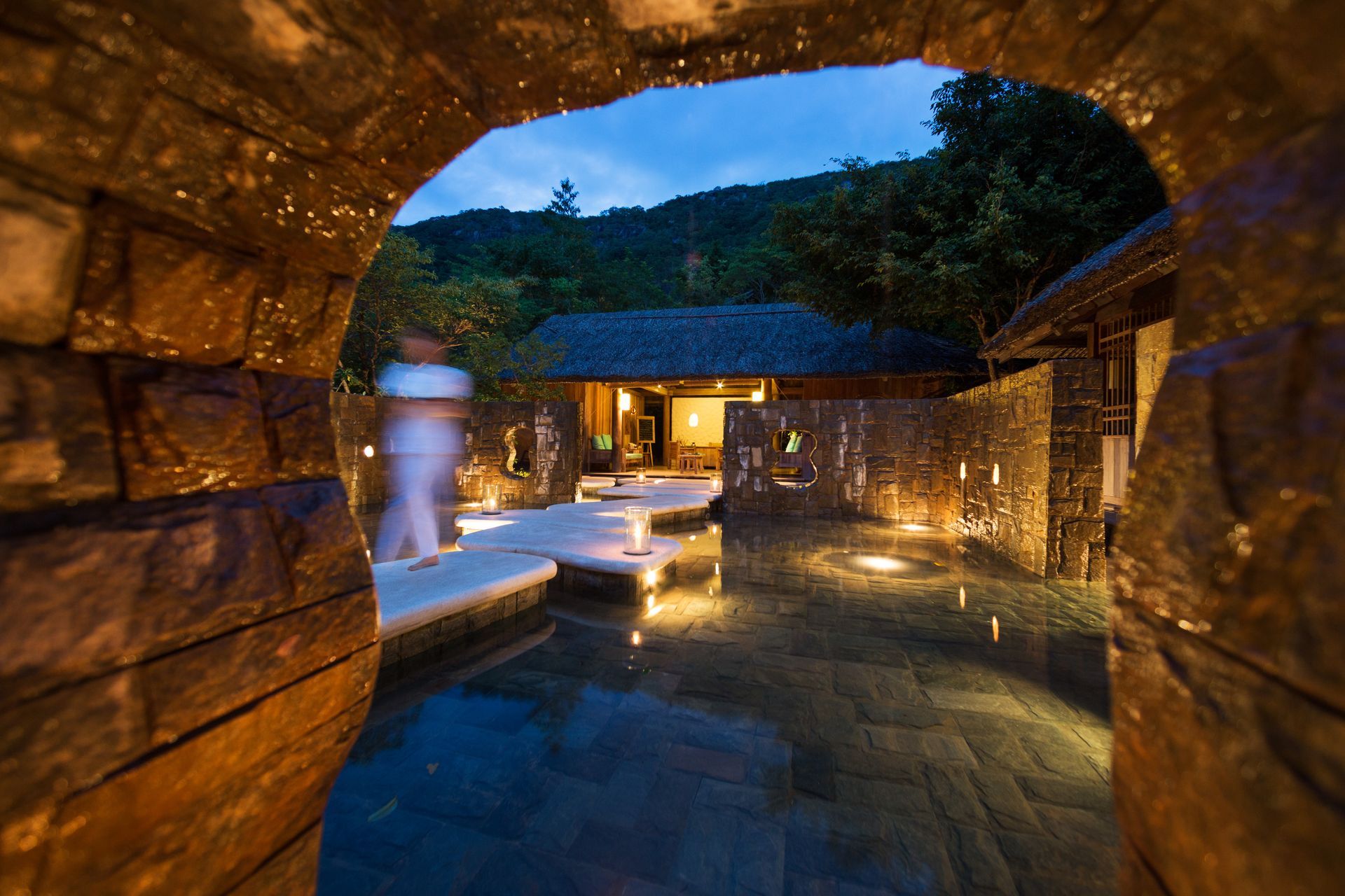 Stone archway frames a spa area with reflecting pool and softly lit buildings at dusk. A person walks by.
