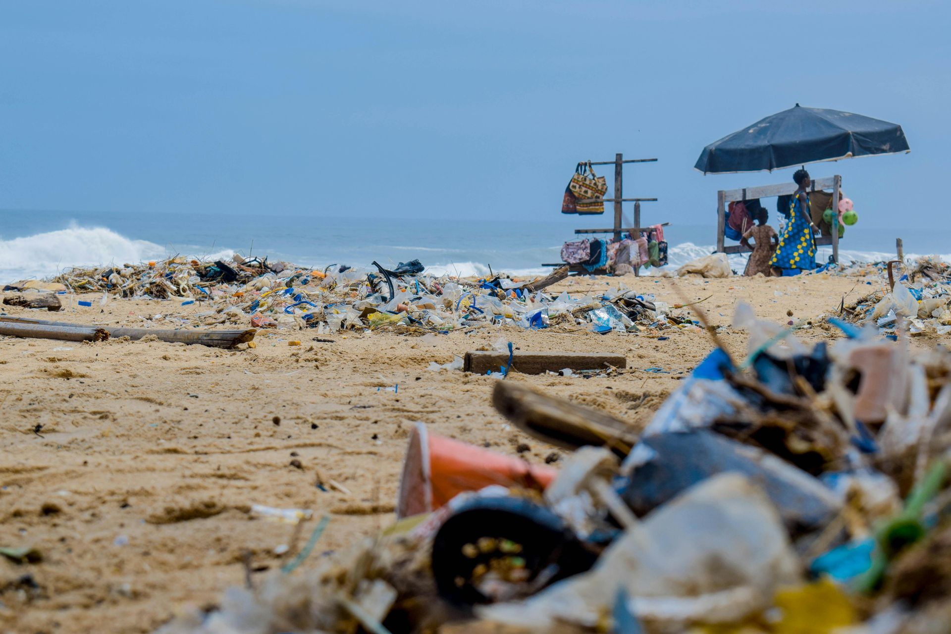 A pile of trash on a beach with vendors sitting in chairs under an umbrella.