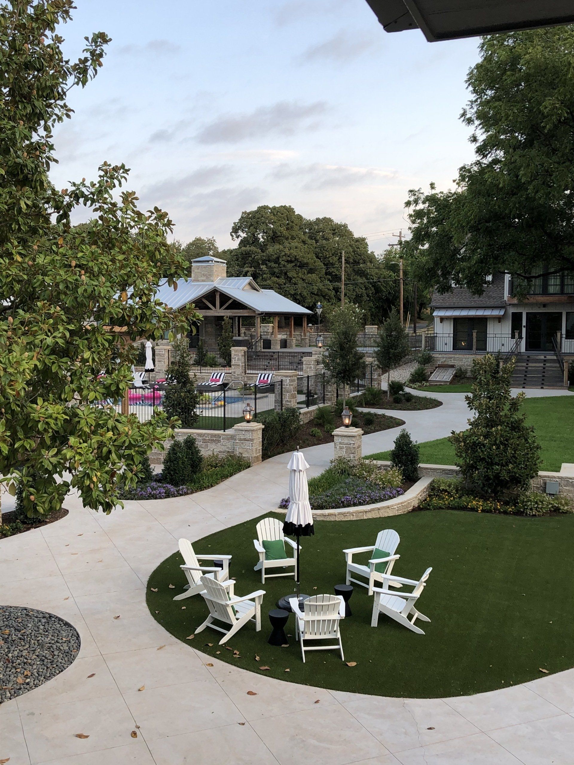 A green lawn with white chairs circles a patio near a large stone building and trees.