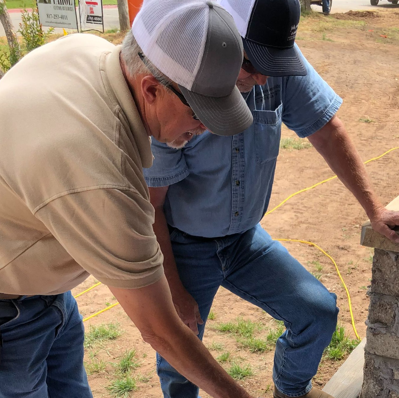 Two men inspecting a stone structure outdoors, both wearing hats, casual clothing.