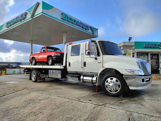 White flatbed tow truck carrying a red pickup under a gas station canopy
