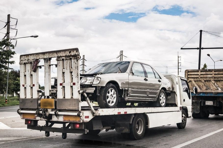 Silver sedan on a tow truck carrying metal cargo, driving on a road under cloudy skies.