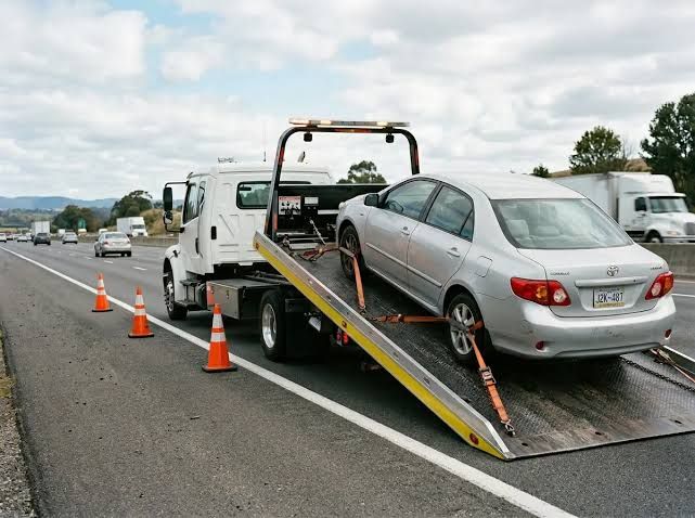 Tow truck loading a silver sedan on a highway shoulder, with orange traffic cones nearby