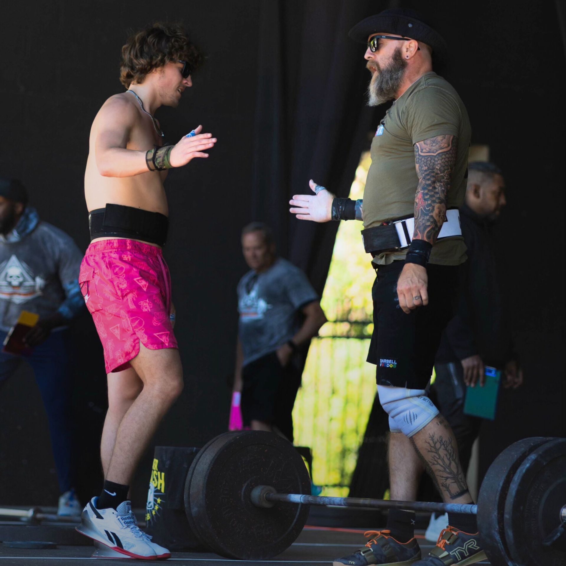 Two men in athletic gear at CrossFit Boiler Room gym  in Villa Rica, GA high-fiving near a barbell. Outdoor setting, sunny.