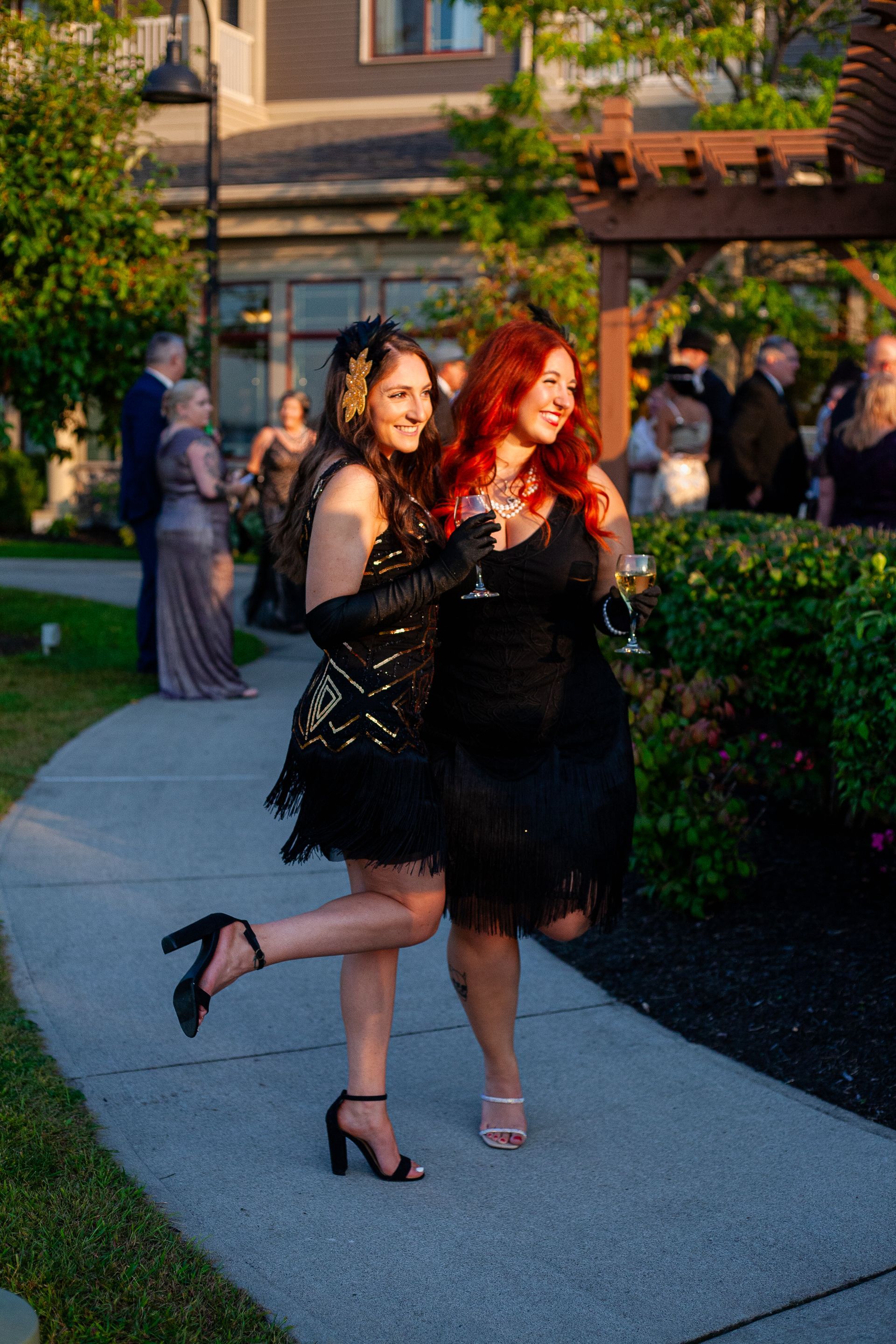 Two women in black dresses are walking down a sidewalk holding wine glasses.