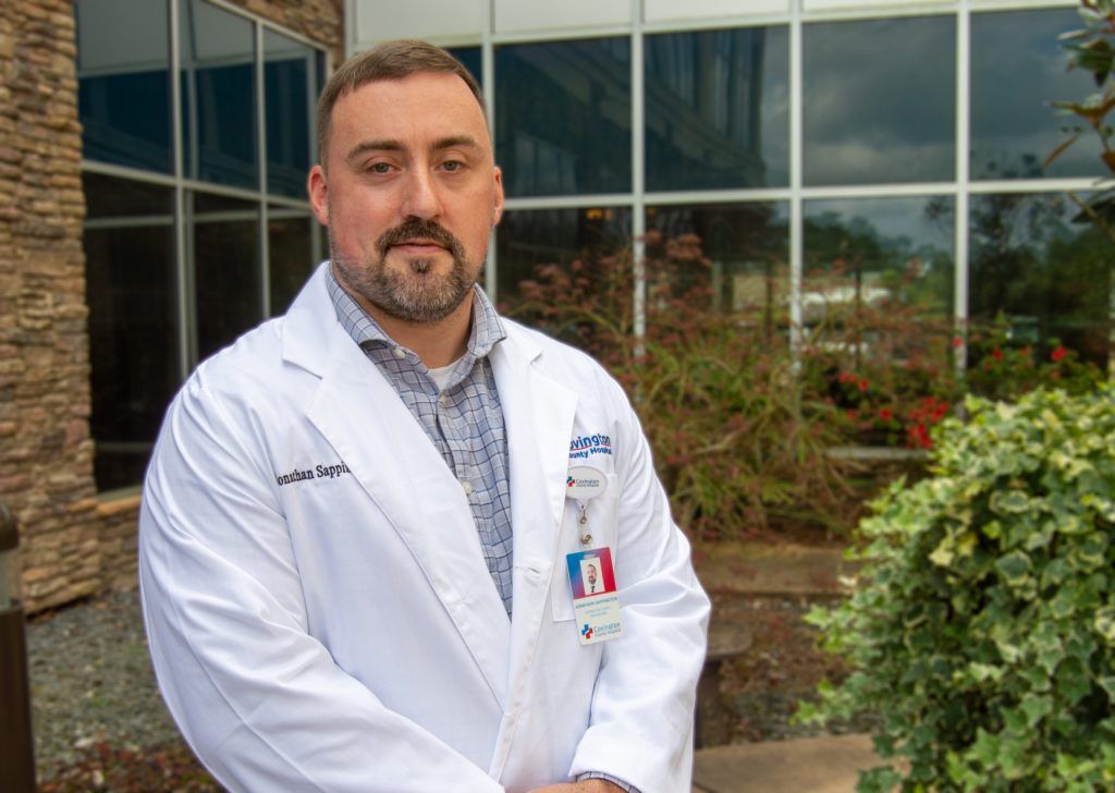 A man in a white lab coat is standing in front of a building.