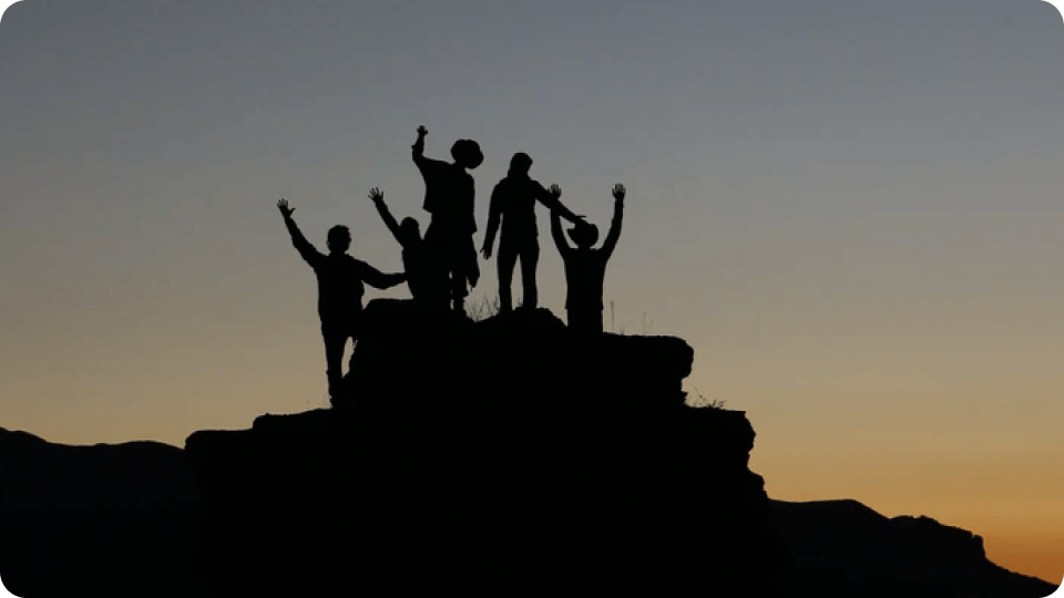 Silhouetted group celebrating on a mountain peak at sunset, arms raised in triumph.