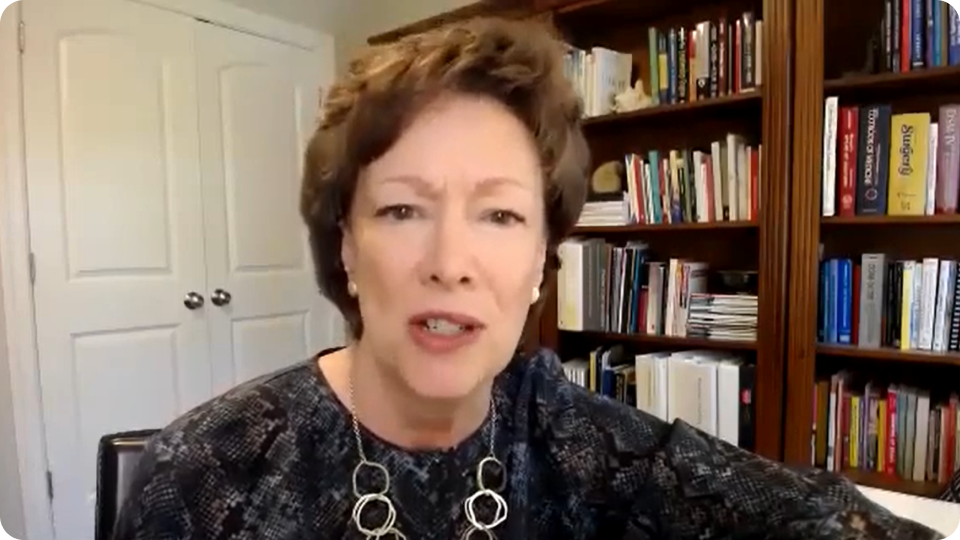 Woman with short brown hair, wearing a patterned blouse, speaking in front of a bookcase.