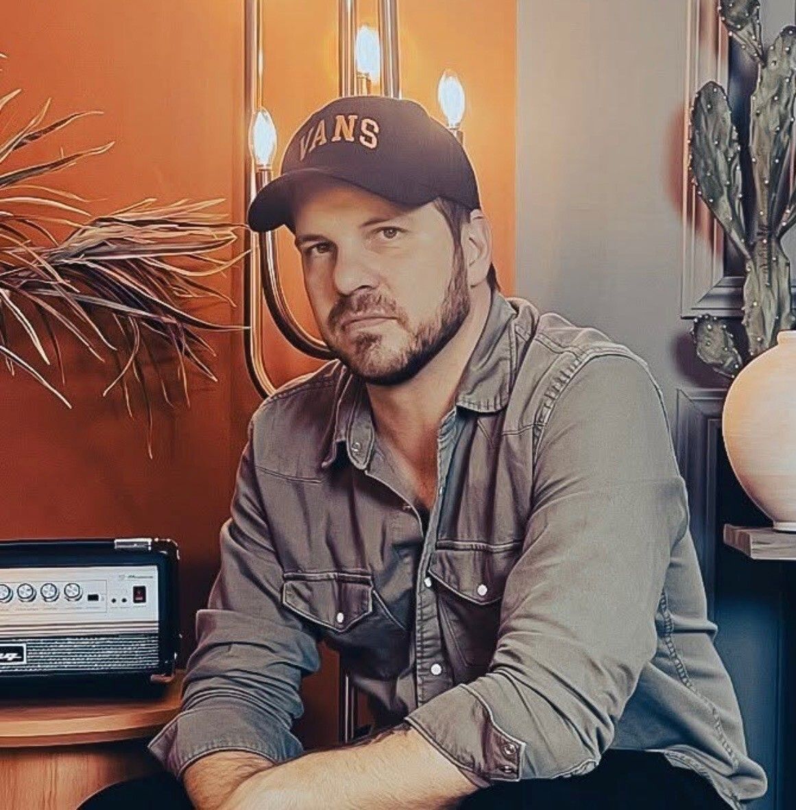 Man seated indoors beside a guitar amplifier, wearing a cap and gray shirt, with warm lighting and decor.