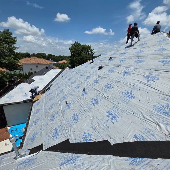 Workers covering a steep roof with white underlayment under a blue sky