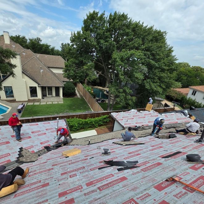Workers installing roofing underlayment on a large roof beside a house and trees