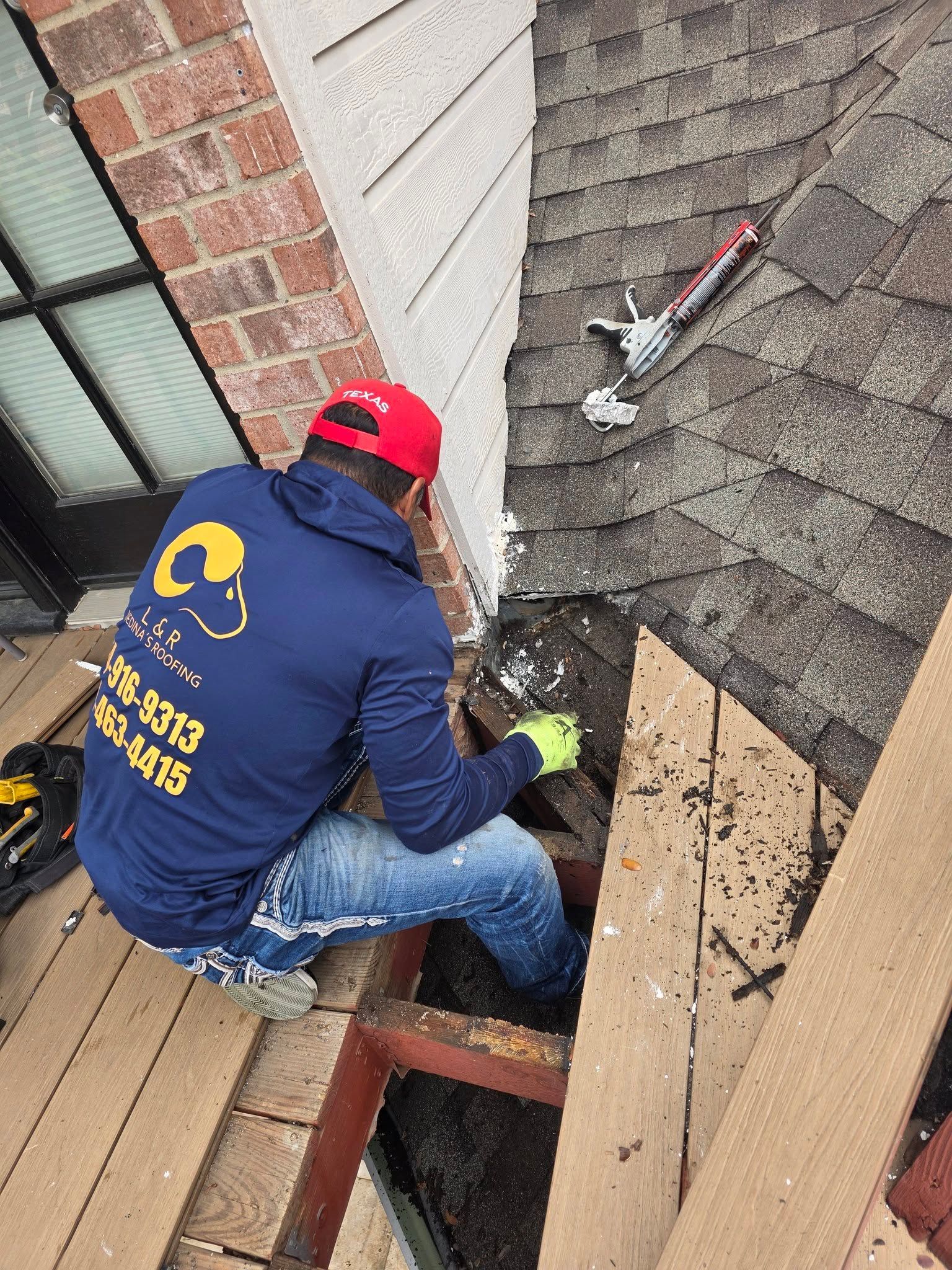 Worker in red cap repairing roof shingles beside a house wall.