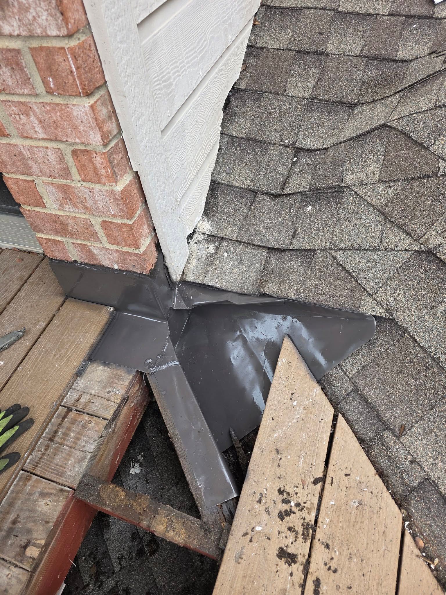 Damaged roof corner with torn shingles, exposed black underlayment, and a broken wooden section near a brick wall