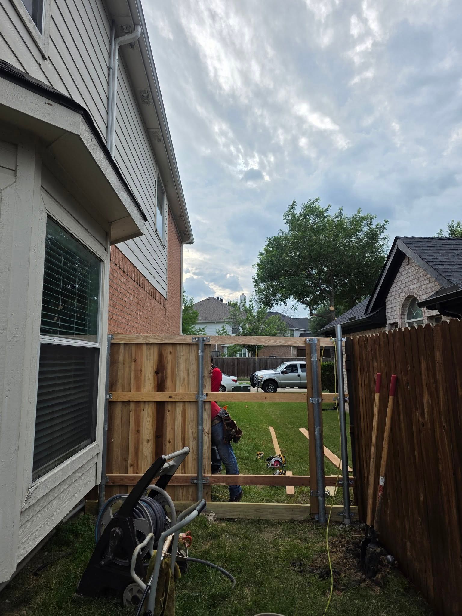 Narrow backyard passage beside a house, with a wooden gate, fence, lawn tools, and cloudy sky.