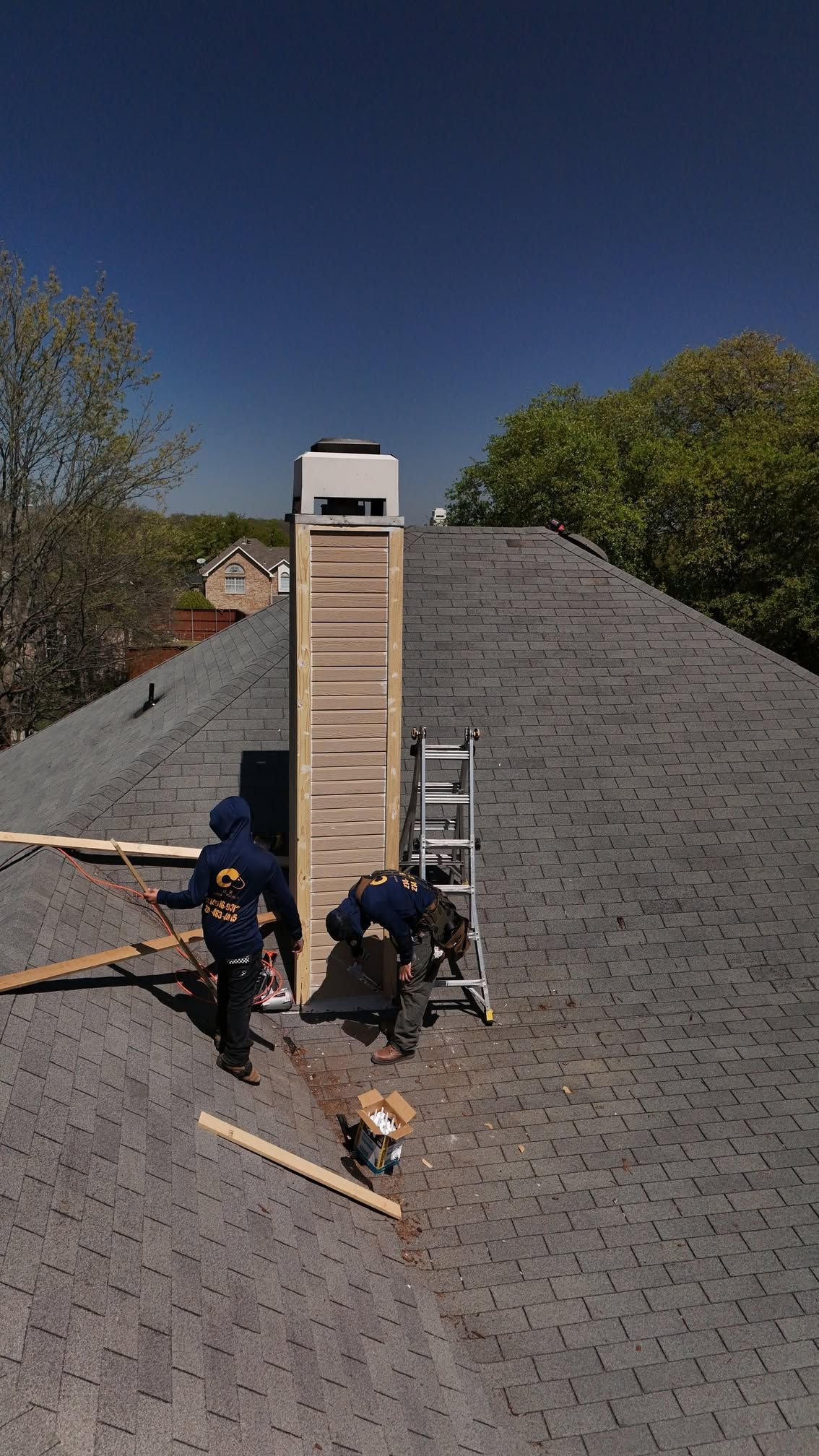Workers repairing a shingled roof beside a tall chimney on a sunny day