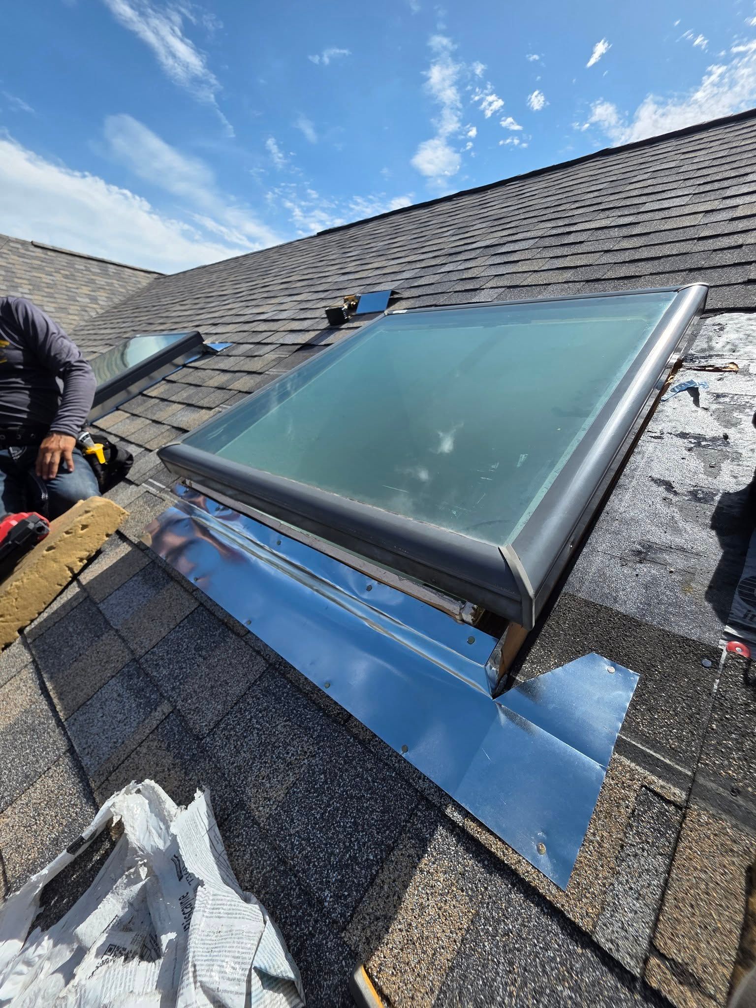 Roof skylight under repair with blue flashing and shingles, worker partially visible on the left