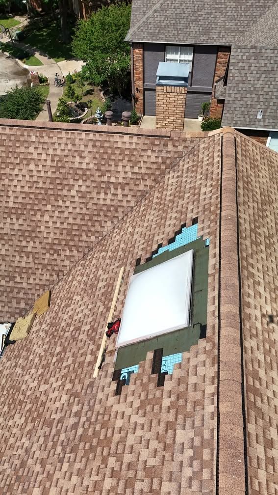 Aerial view of a shingled roof with a skylight beside a brick house and garden below.