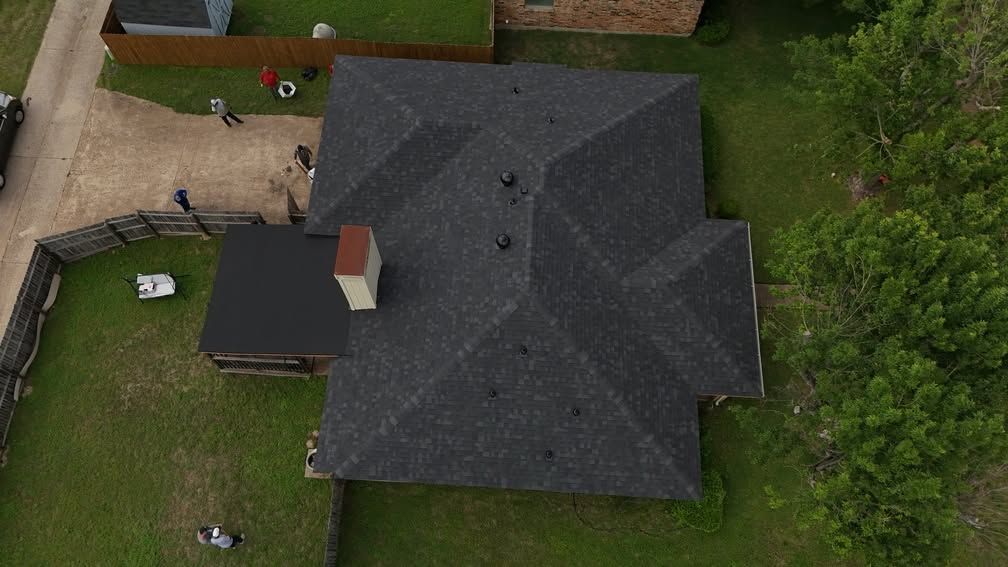 Aerial view of a house with a dark roof, fenced yard, and surrounding trees.