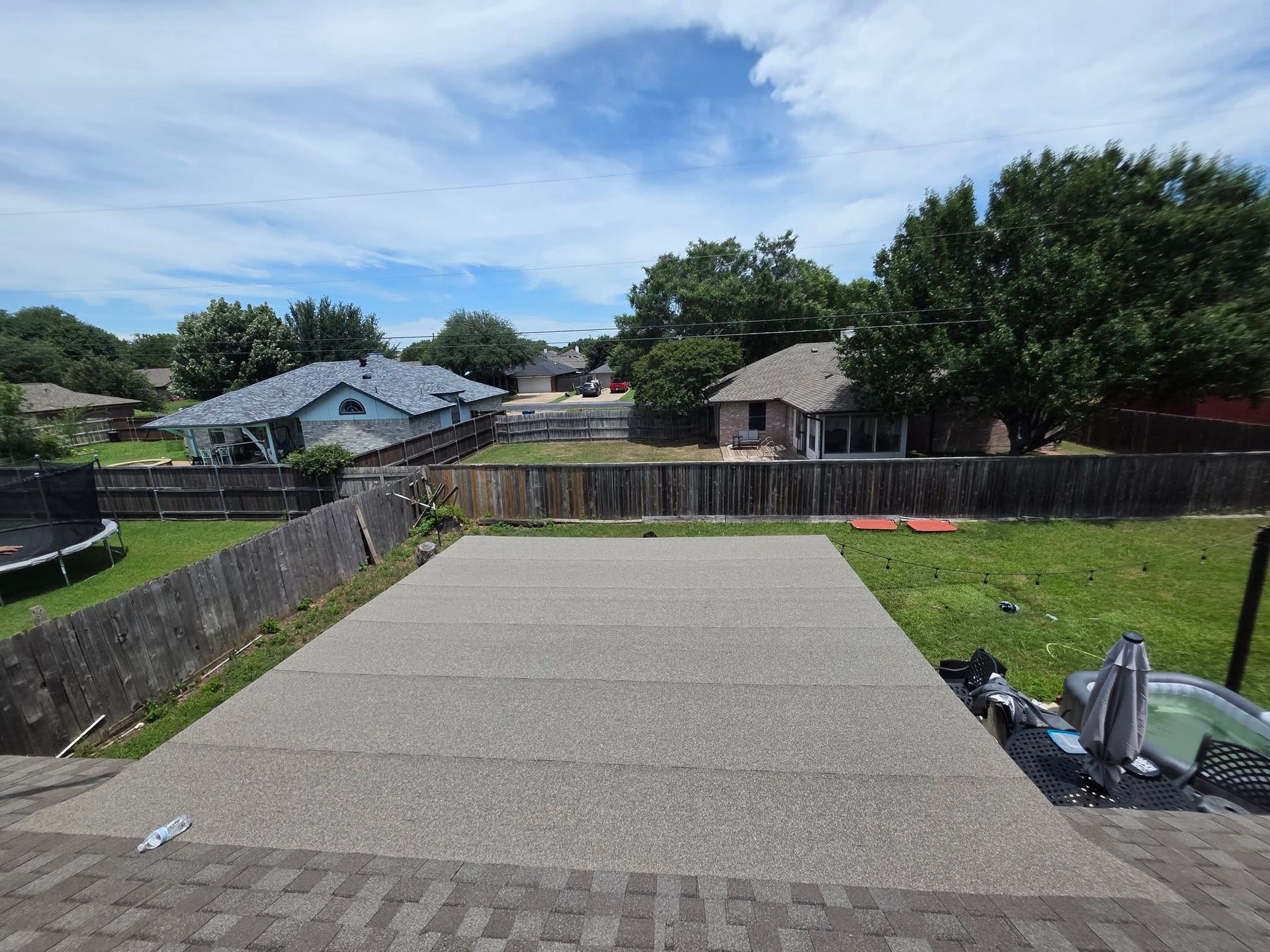 Backyard patio with a concrete slab, grass, wooden fences, and neighboring houses under a blue sky