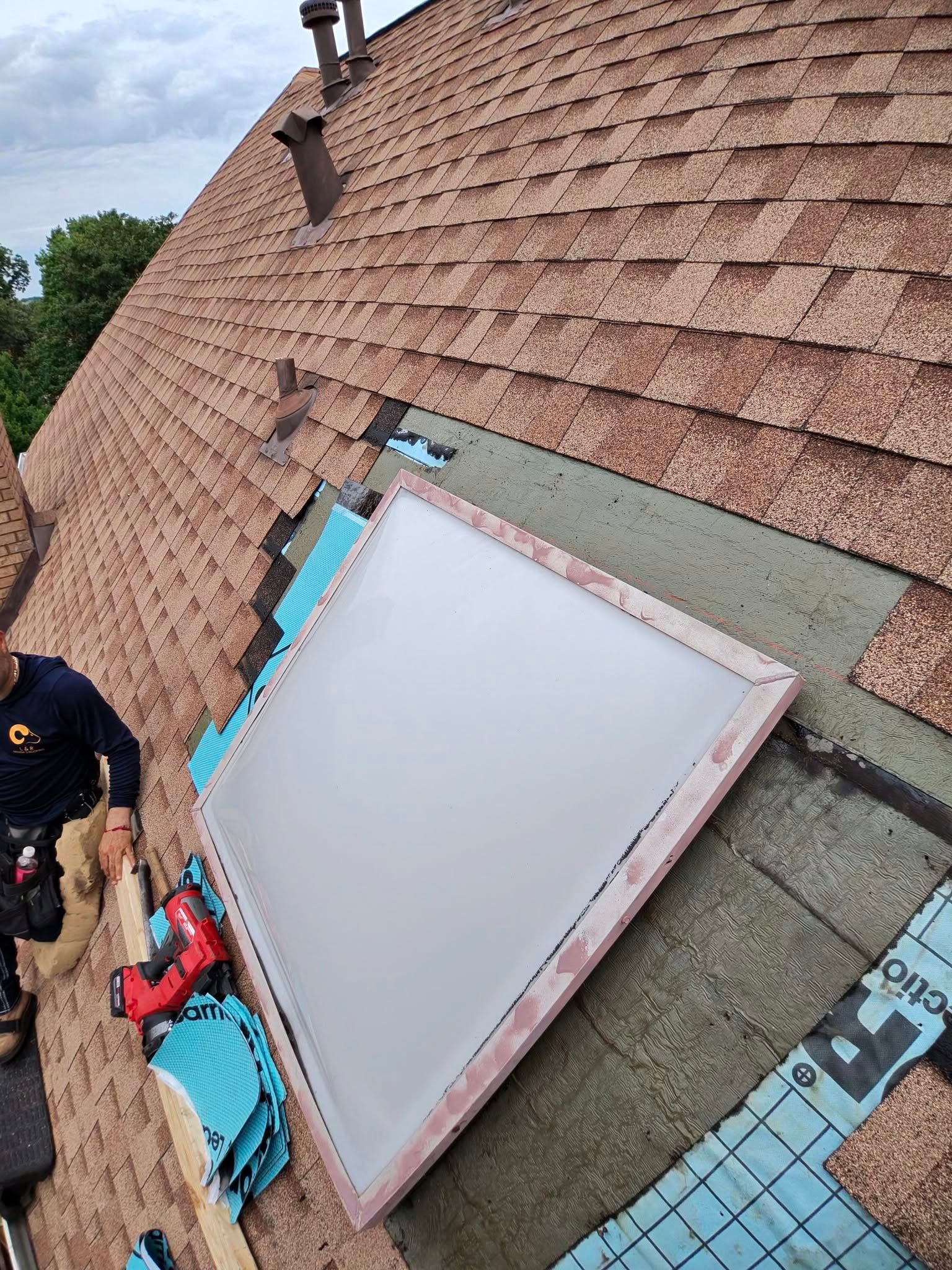 Roofers working around a skylight on a shingled roof, with tools and materials nearby.