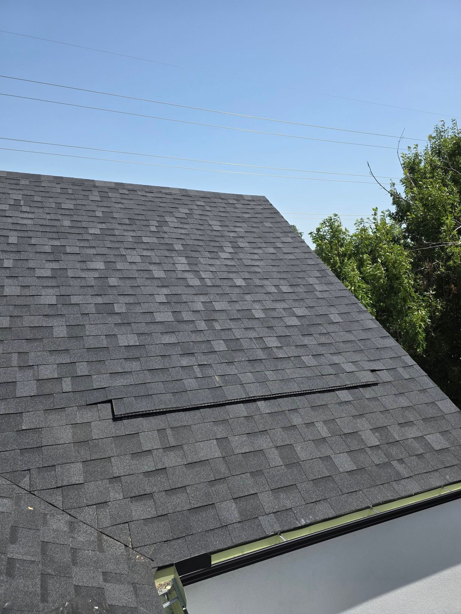 Gray shingled roof with a partially lifted lower row, against a blue sky and trees
