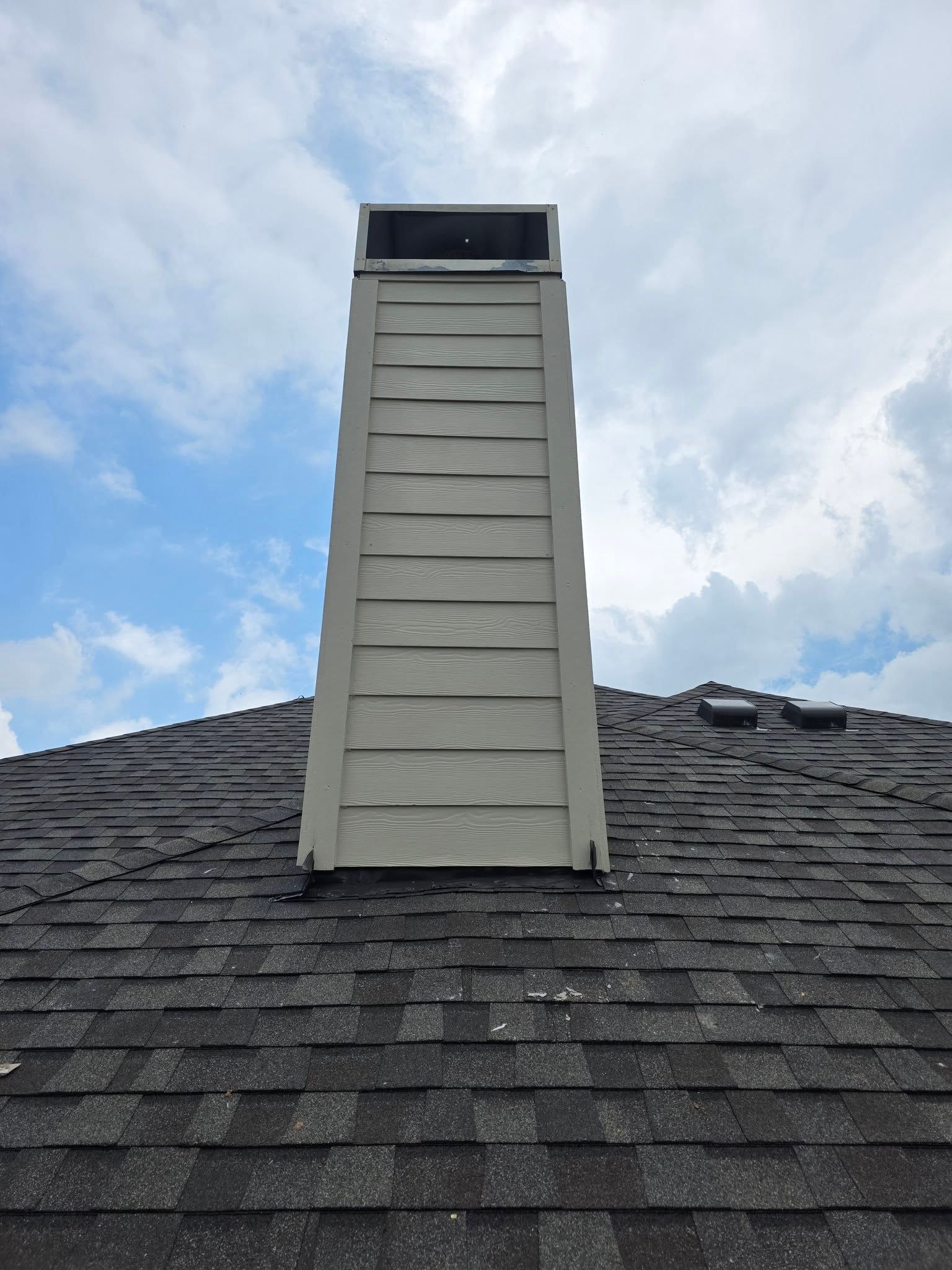 Tall beige roof chimney with dark cap on a gray shingled roof against a cloudy sky