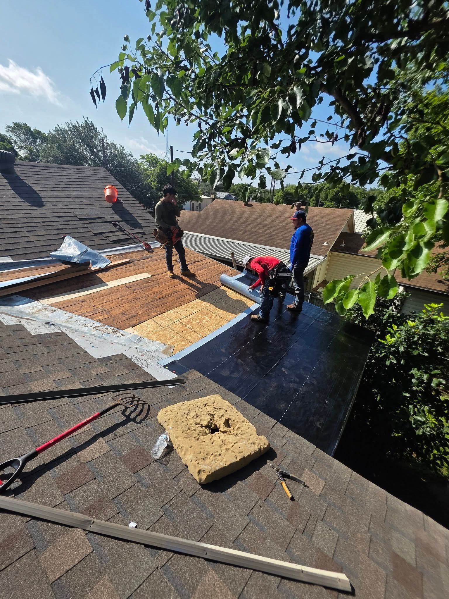 Roofers installing tar paper on a sloped roof under a tree, with tools and materials nearby.
